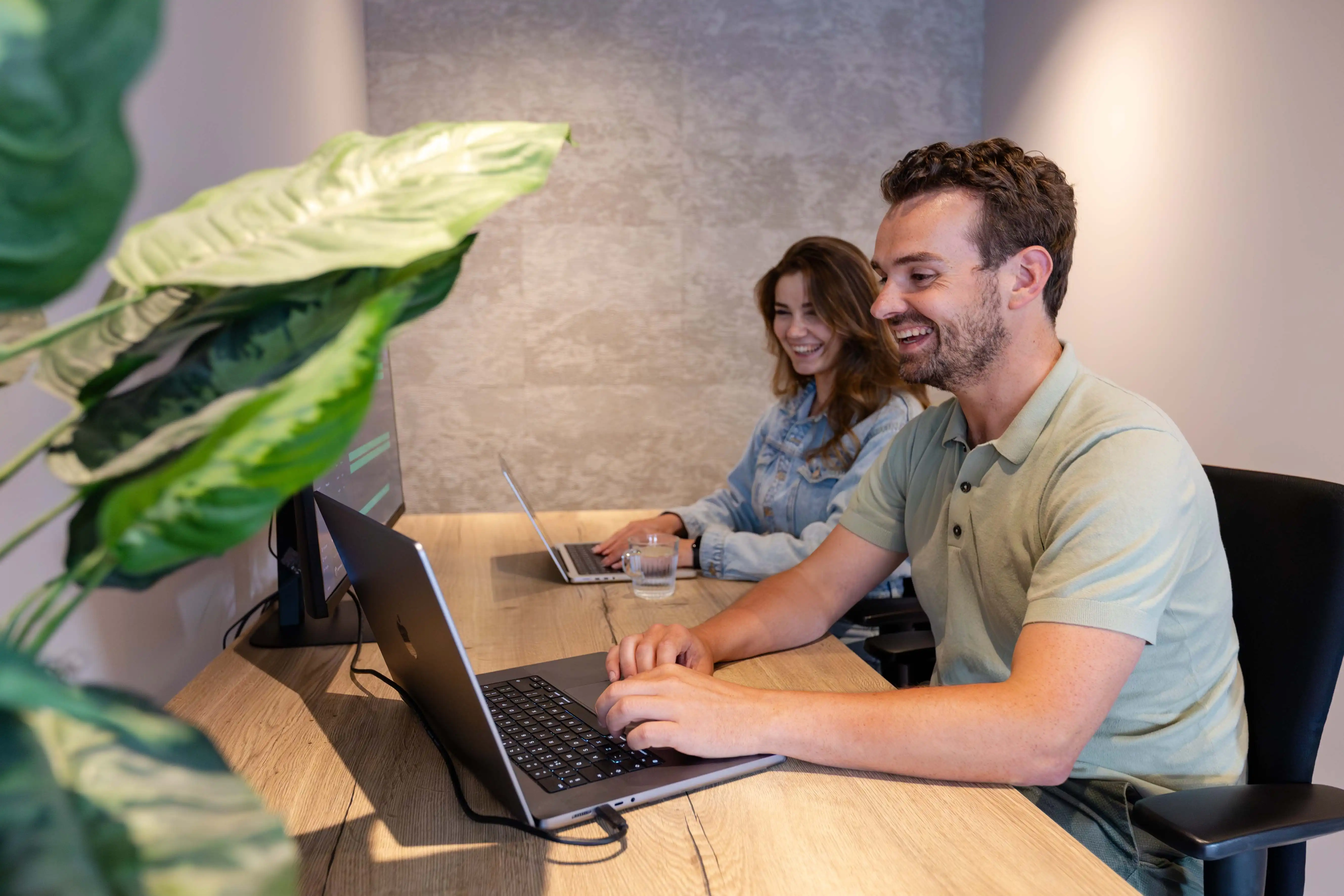 Een man en een vrouw met bruin haar van planning software bedrijf vPlan zitten lachend achter hun laptop aan een bureau.